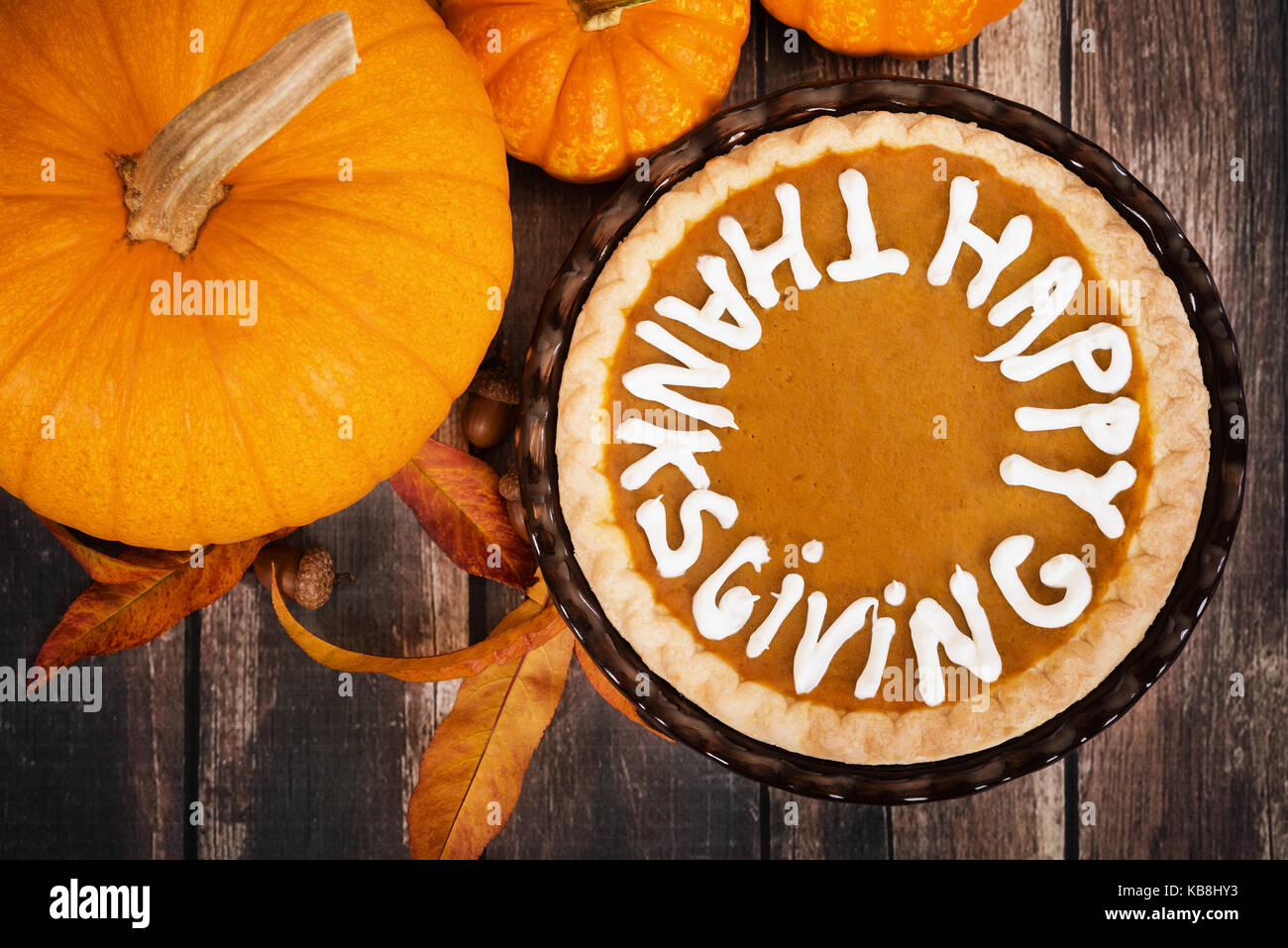 Pumpkin pie with Happy Thanksgiving text. Displayed with pumpkins, golden autumn leaves, and acorns on vintage, wooden table. Top view. Stock Photo