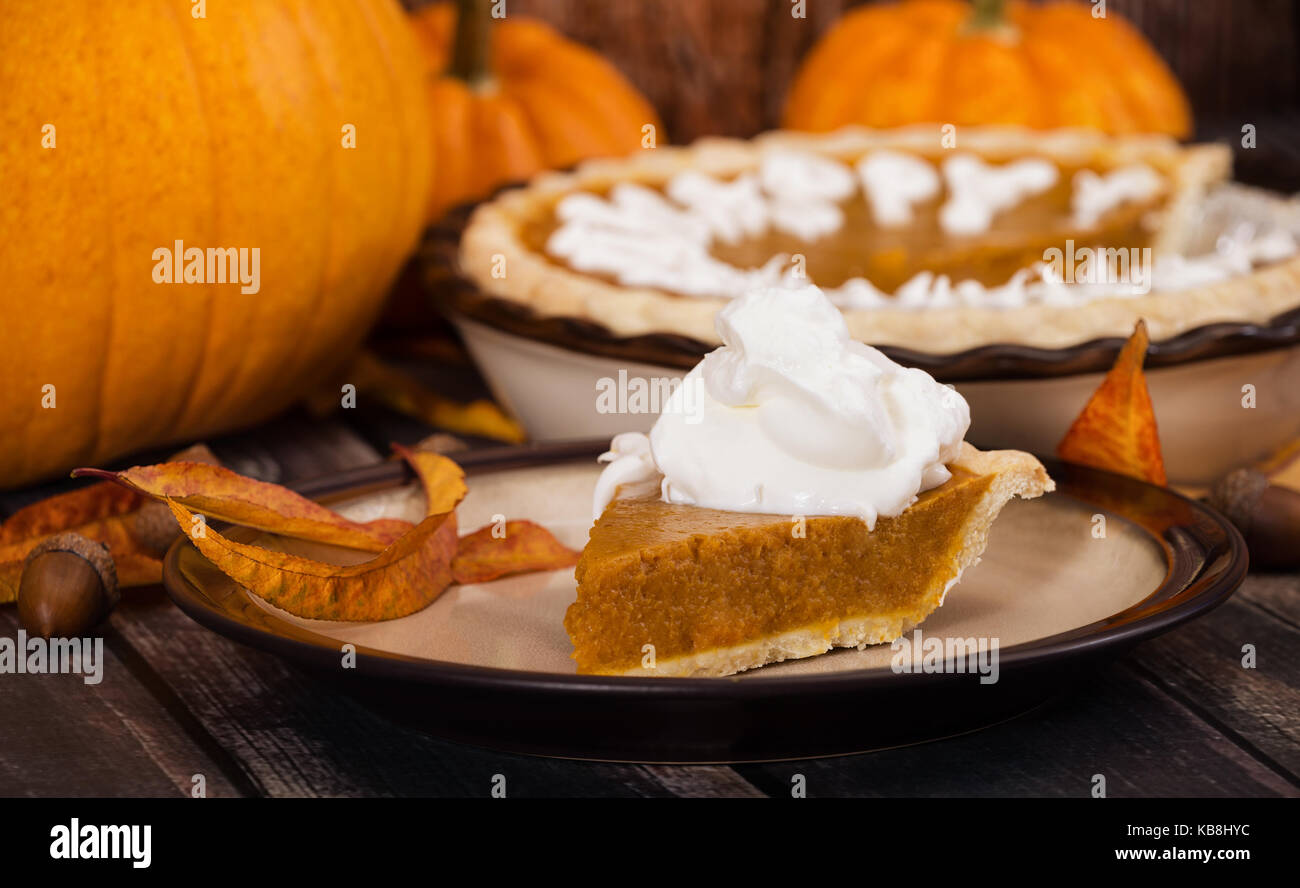 A slice of a pumpkin pie served with whipped cream. Pumpkins and autumn decorations diplayed on rustic table. Stock Photo