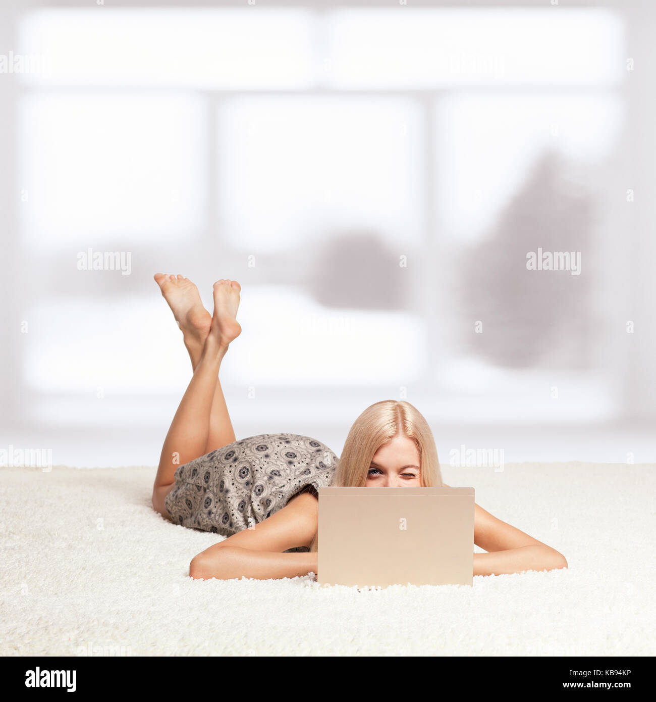 Winking blonde young woman on white whole-floor carpet browsing laptop  near window Stock Photo
