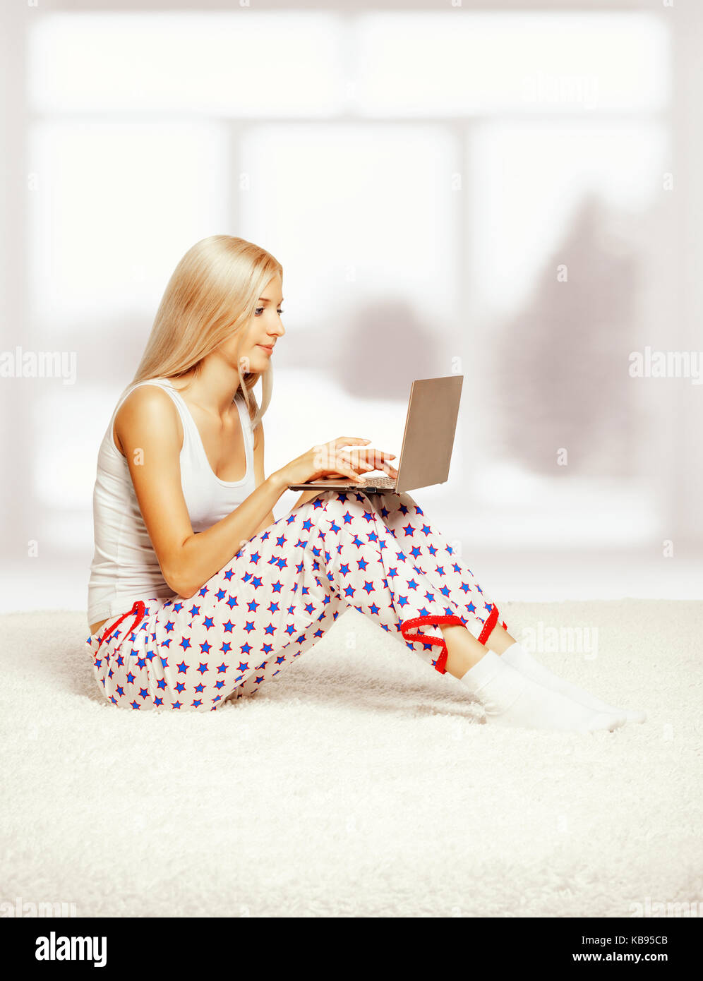 Young blonde woman in pyjamas on white whole-floor carpet browsing laptop  near window Stock Photo