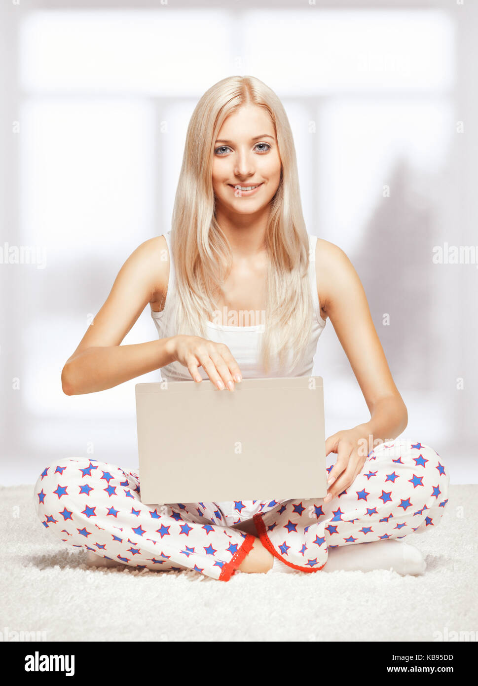 Young blonde woman in pyjamas sitting on white whole-floor carpet browsing laptop  near window Stock Photo