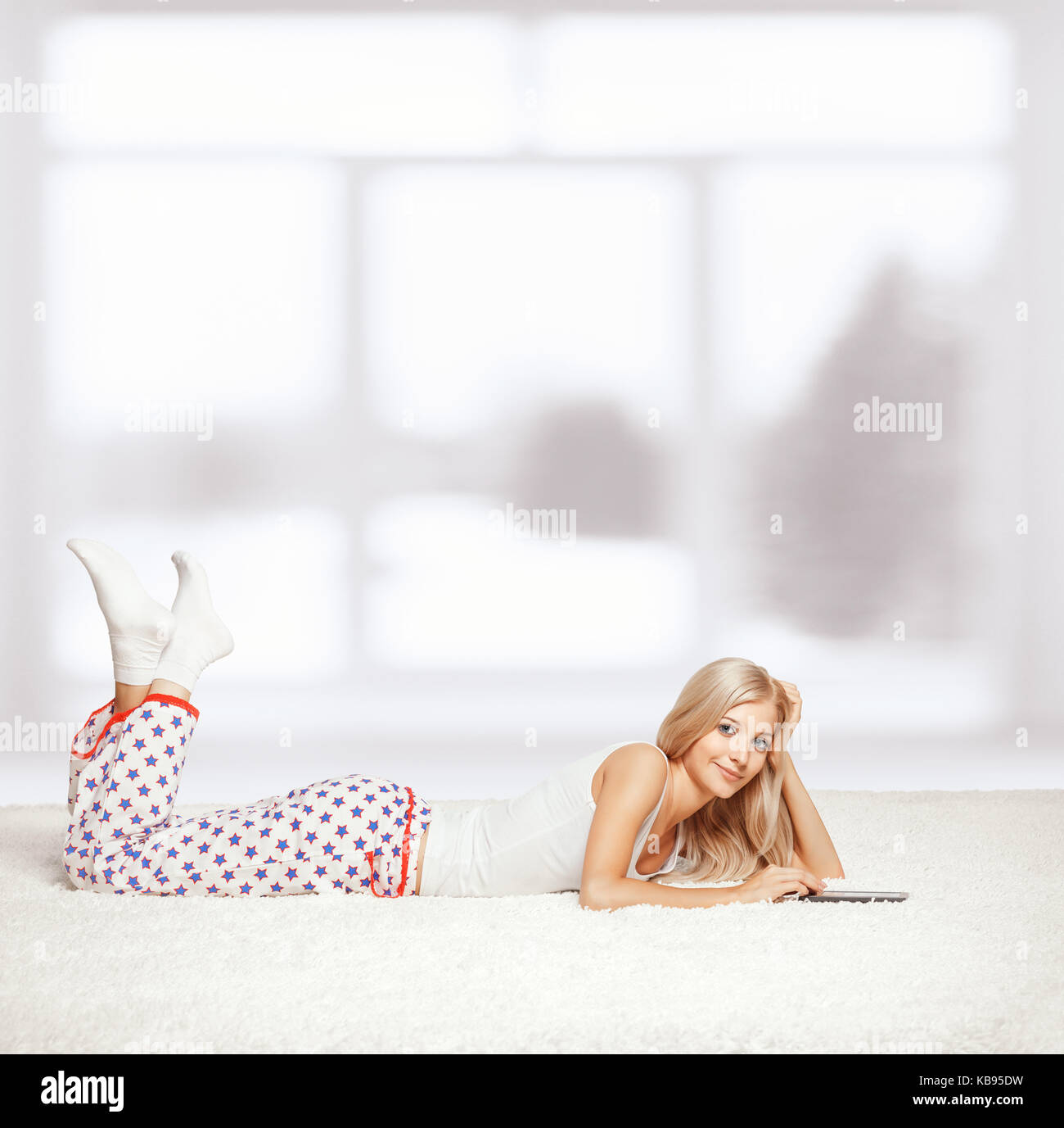 Young blonde woman in pyjamas on white whole-floor carpet reading e-book Stock Photo