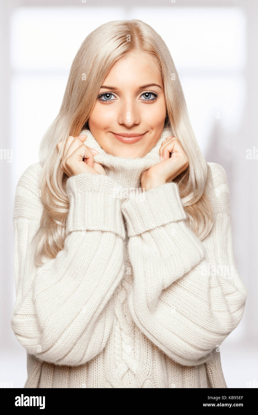 Blonde young beautiful woman dressed in large white cashmere sweater on window background Stock Photo