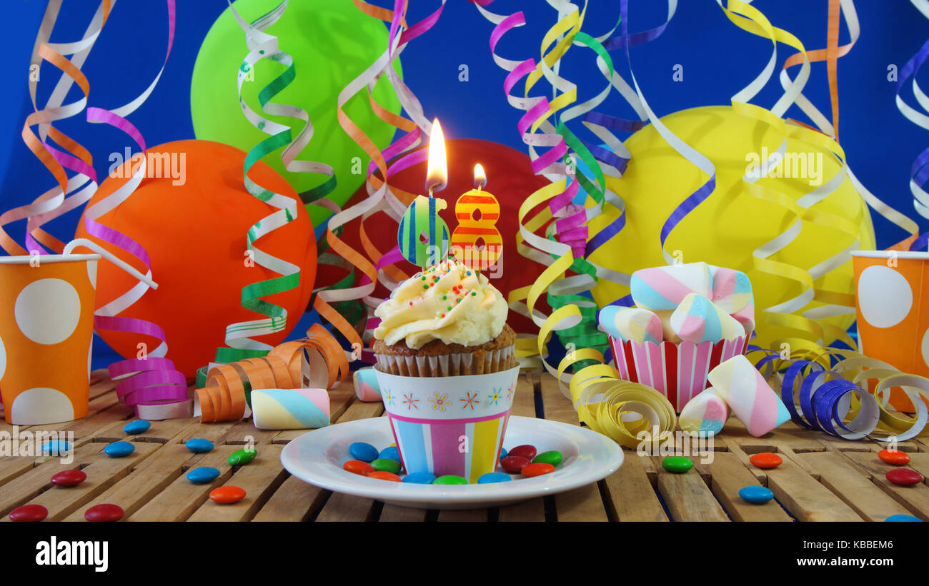 68 Birthday cupcake with candles burning on rustic wooden table with background of colorful balloons plastic cups candies on blue wall in background Stock Photo
