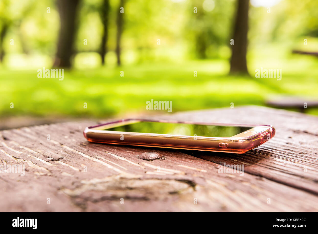 Macro closeup of lost generic light pink rose gold smartphone, phone left, forgotten in plastic clear case lying on table in outside summer park Stock Photo