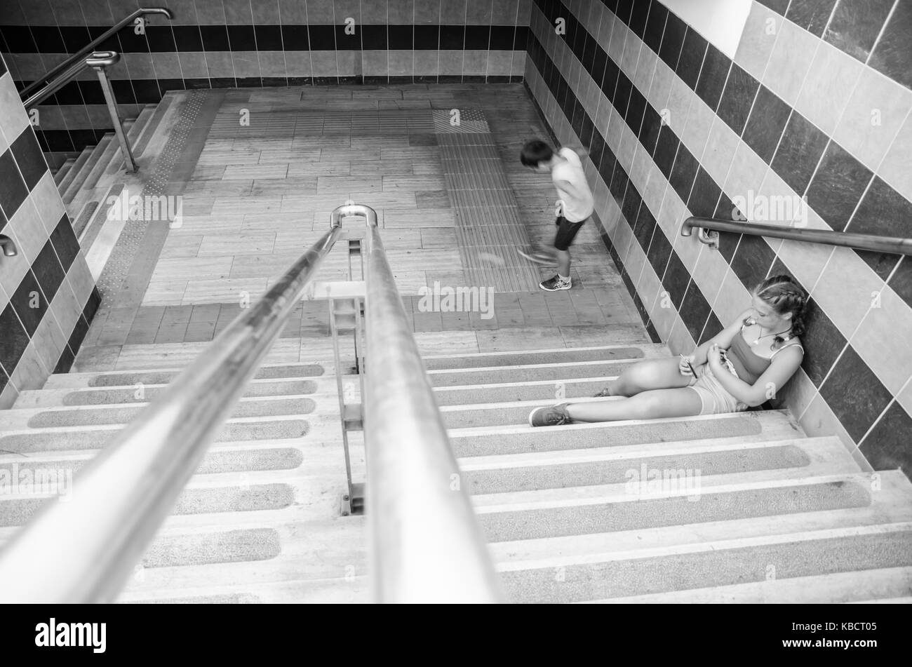 Waiting the train. Caucasian girl in shorts and t-shirts sitting on stairs of a railway station while consulting her smartphone. Caucasian boy is play Stock Photo