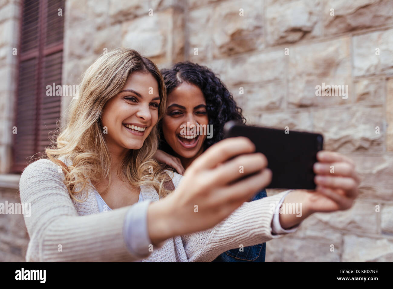 cheerful girls taking a selfie using mobile phone women standing outdoors KBD7NE