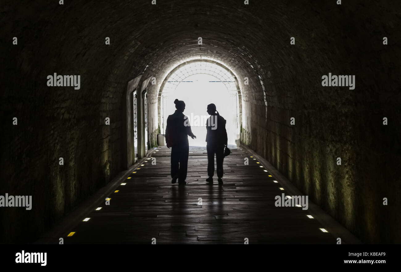 Two girls walking out of a dark corridor made of stone, towards the bright light of the day. Stock Photo