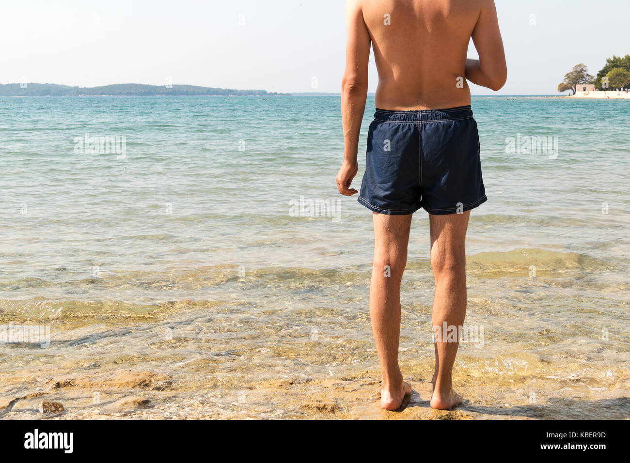 Masculine model of backs looking at the sea. Clear water, bright sun and sunscreen for sunburn. Man in blue shorts getting ready to go swimming in the Stock Photo