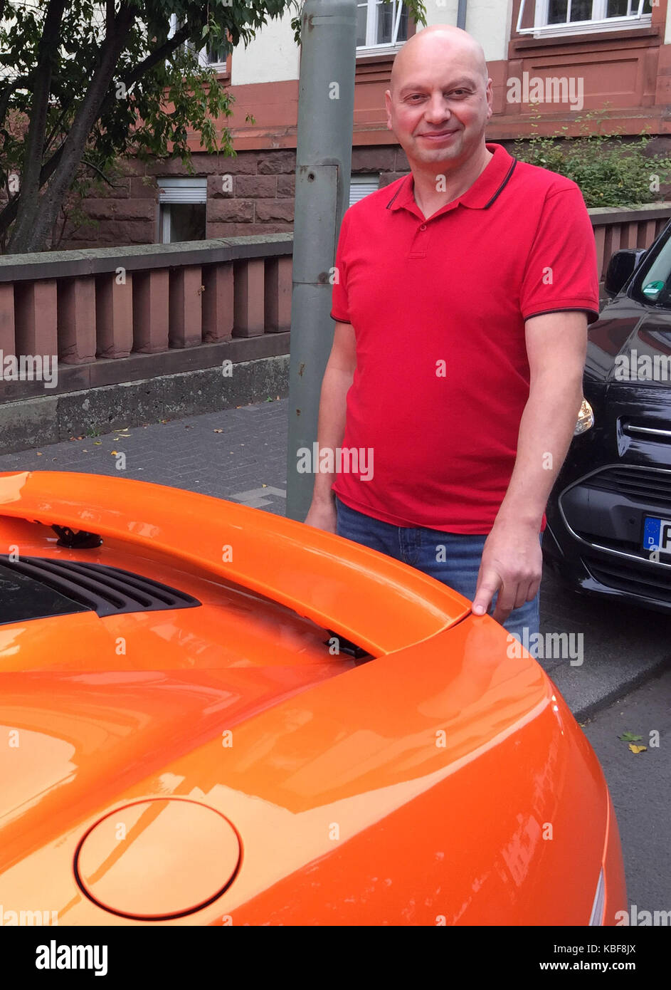 Markus Zahn Stands Behind His Orange Sports Car And Points Where A 