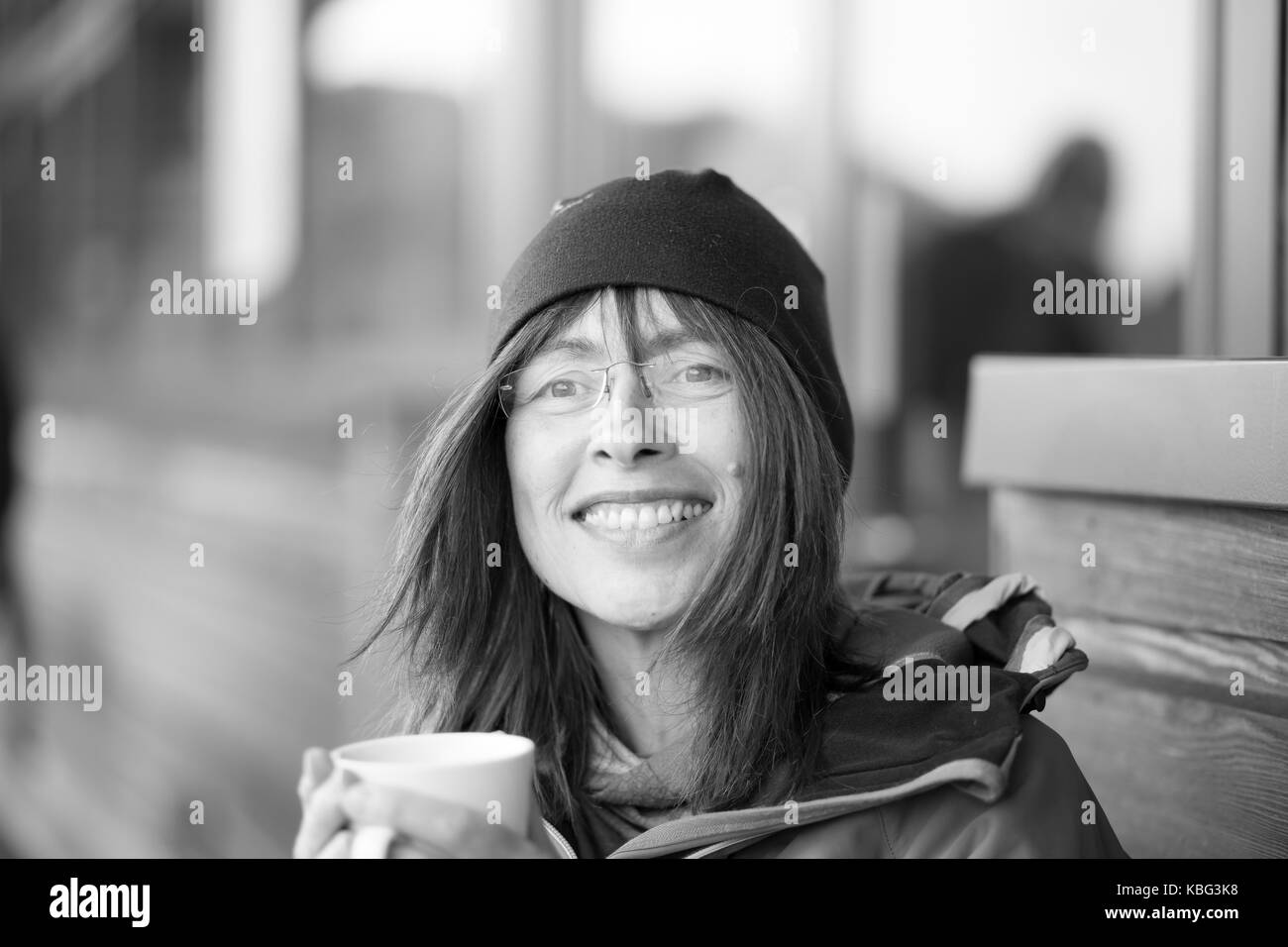 Black & white close up portrait of trendy 50 year old, young outlook, in beanie hat isolated outdoors holding coffee cup with big, natural smile. Stock Photo