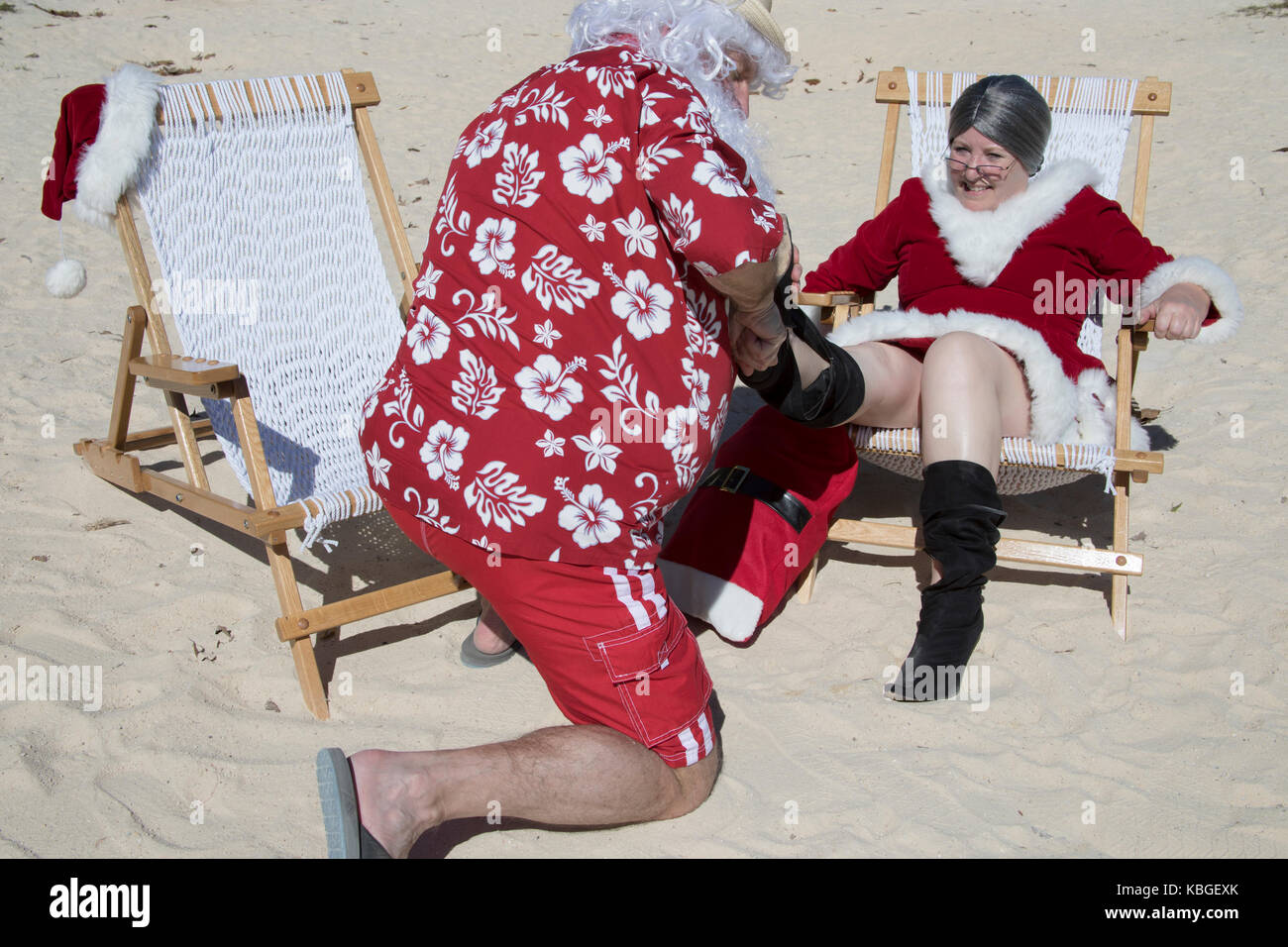 Santa Claus in red swimming trunks and Hawaiian shirt  on sandy beach helping to remove Mrs Claus boots. Stock Photo
