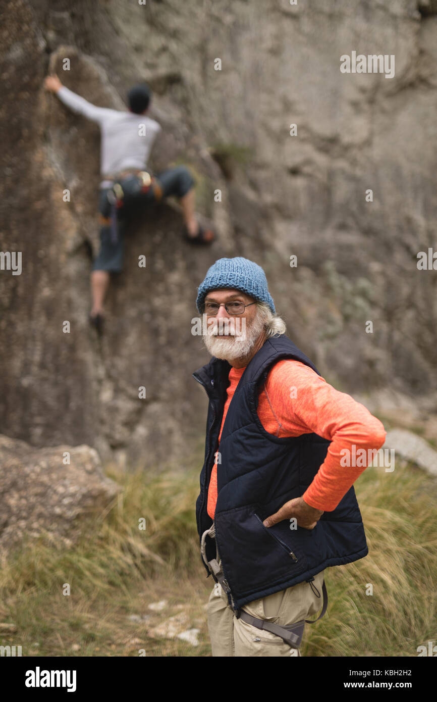 Portrait of senior man standing with hand on hip while friend climbing rock in background Stock Photo