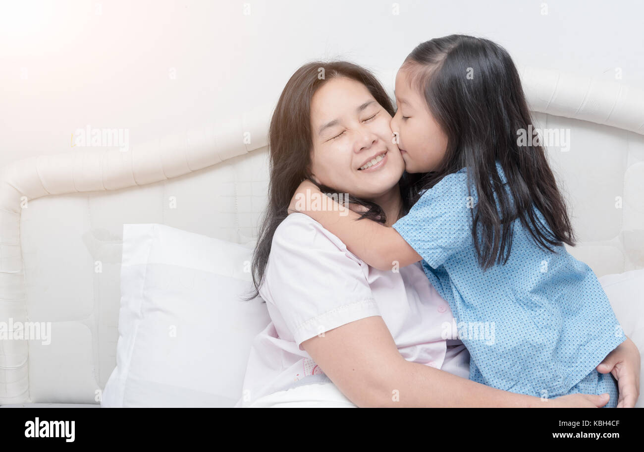 Asian sweet little girl is hugging and kissing her beautiful young mom in cheek while sitting on bed at home, love concept Stock Photo