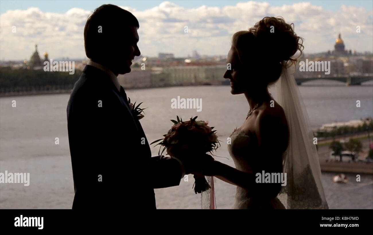 Bride and groom stand smiling in a front of a window hidden by golden ...