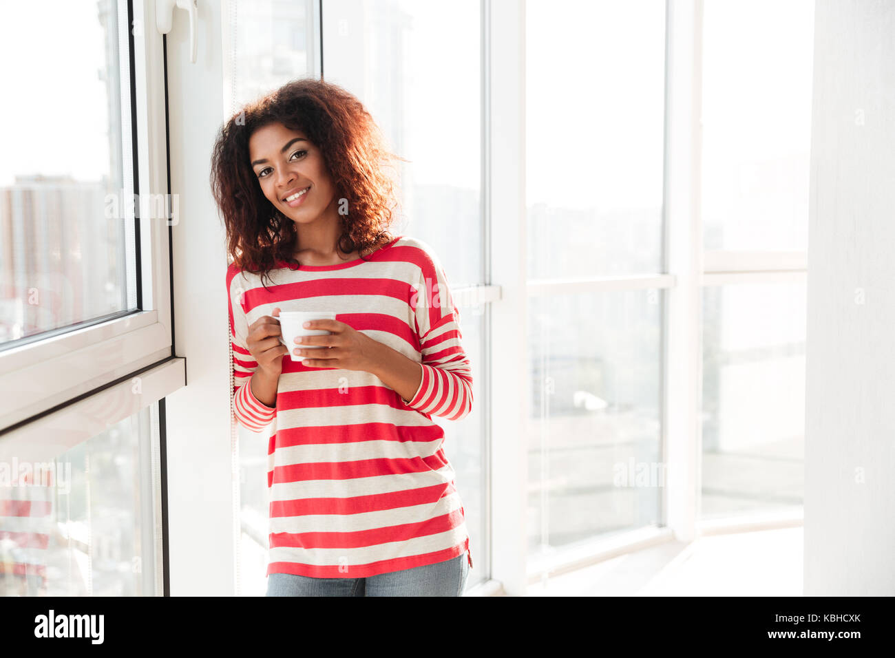 Happy african woman drinking coffee near the window and looking at the ...