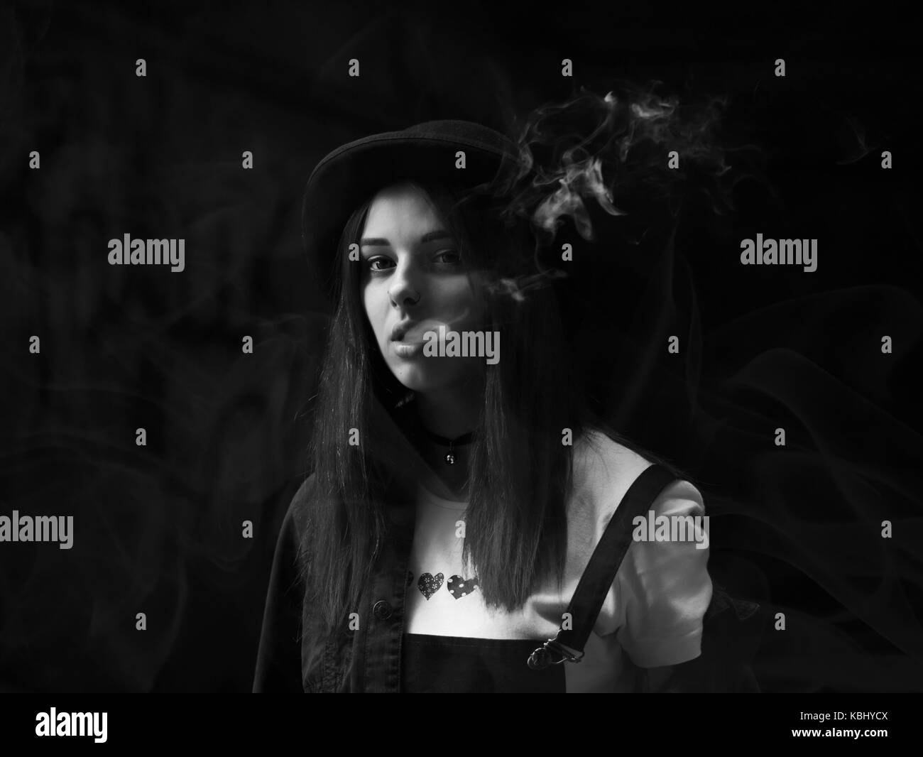 Emo girl smoking cigarette. Young student or pupil with blue colorful dyed  hair, hat, piercing,lenses,ears tunnels and unusual hairstyle stands on  black background. Photos
