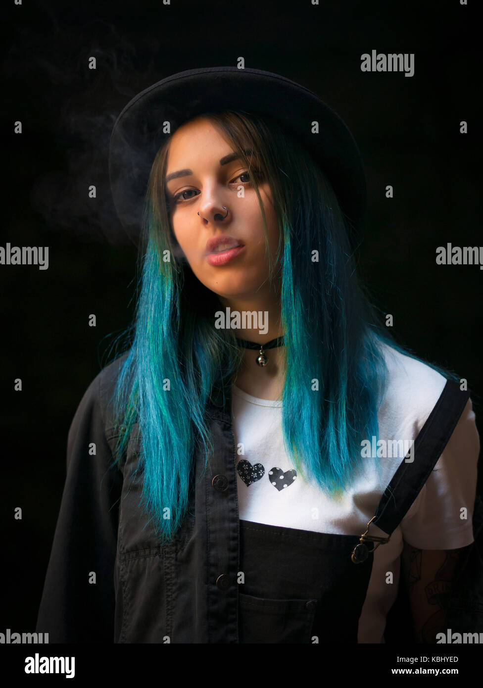 Emo girl smoking cigarette.Street punk or hipster woman with blue colorful dyed hair, hat, piercing,lenses,ears tunnels and unusual hairstyle stands i Stock Photo