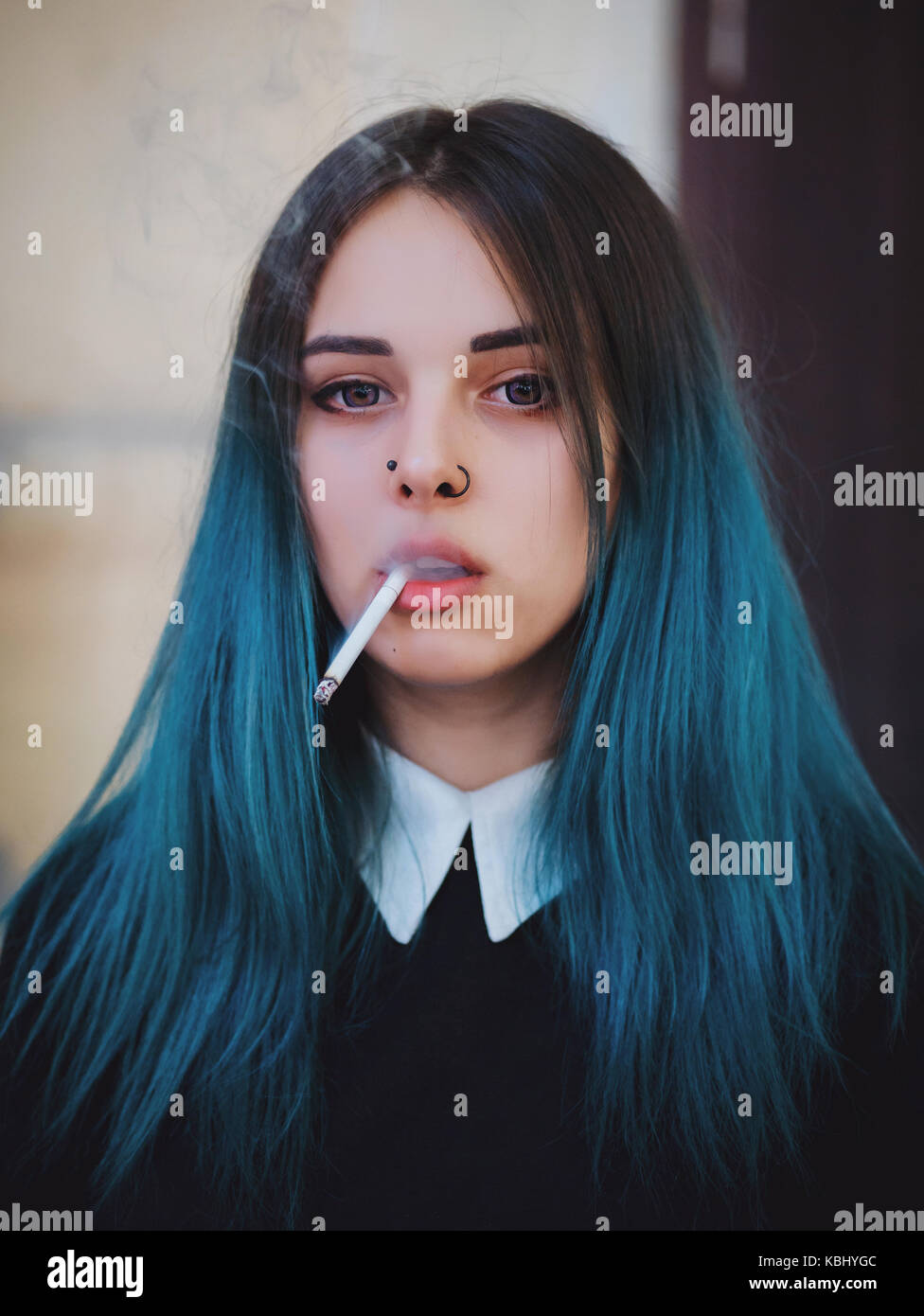 Emo girl smoking cigarette. Young student or pupil with blue colorful dyed hair, hat, piercing,lenses,ears tunnels and unusual hairstyle stands on bla Stock Photo