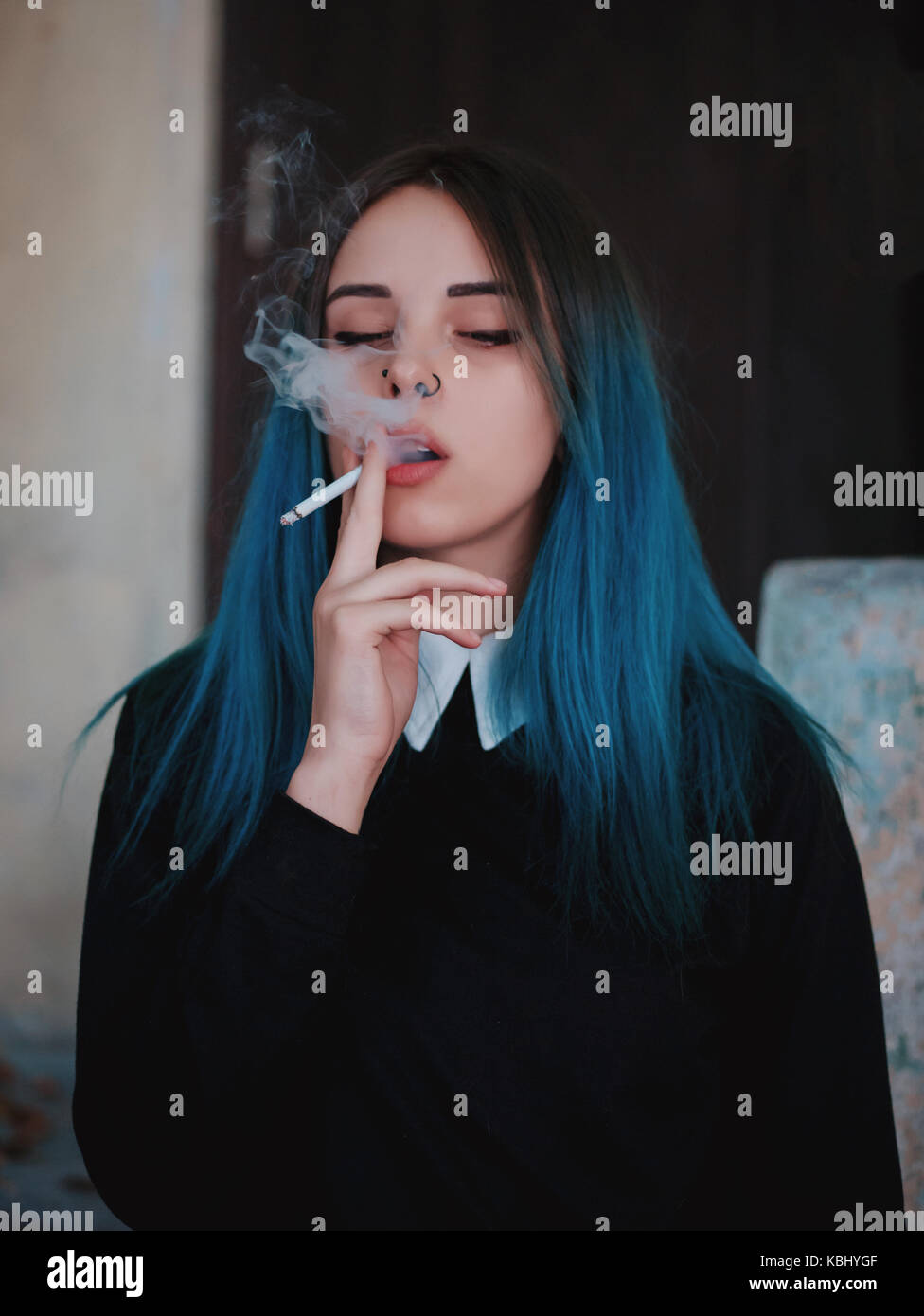 Emo girl smoking cigarette. Young student or pupil with blue colorful dyed hair, hat, piercing,lenses,ears tunnels and unusual hairstyle stands on bla Stock Photo