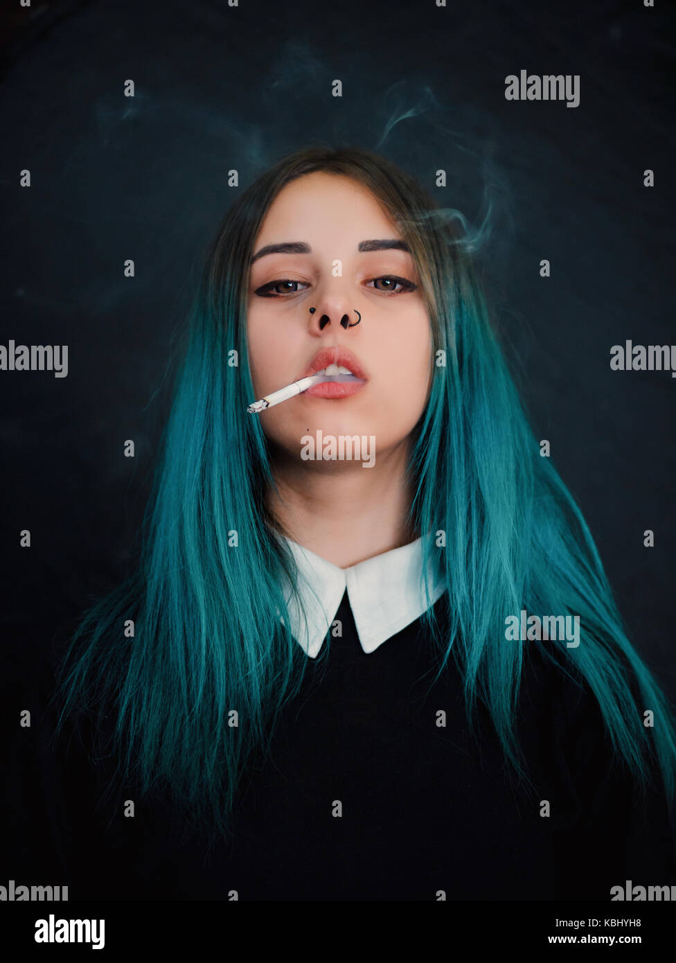 Emo girl smoking cigarette. Young student or pupil with blue colorful dyed hair, hat, piercing,lenses,ears tunnels and unusual hairstyle stands on bla Stock Photo