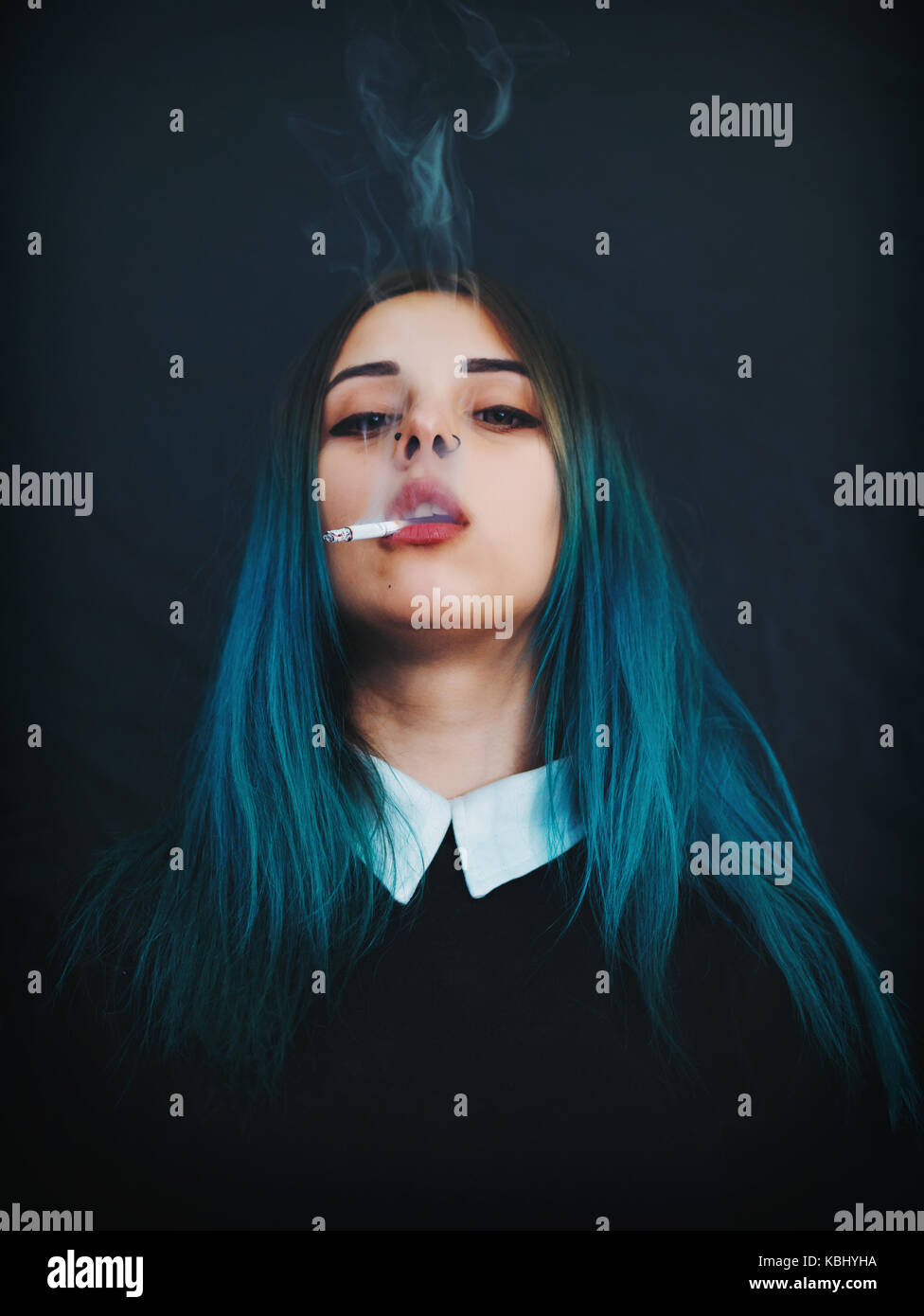 Emo girl smoking cigarette. Young student or pupil with blue colorful dyed hair, hat, piercing,lenses,ears tunnels and unusual hairstyle stands on bla Stock Photo