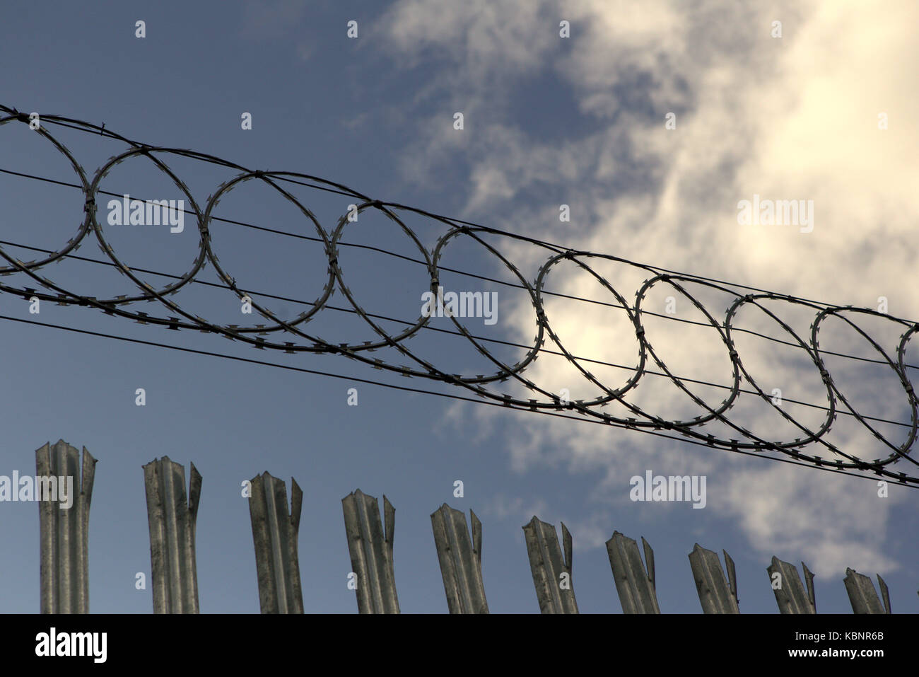 razor wire barbed wire steel fence top blue cloudy sky Stock Photo