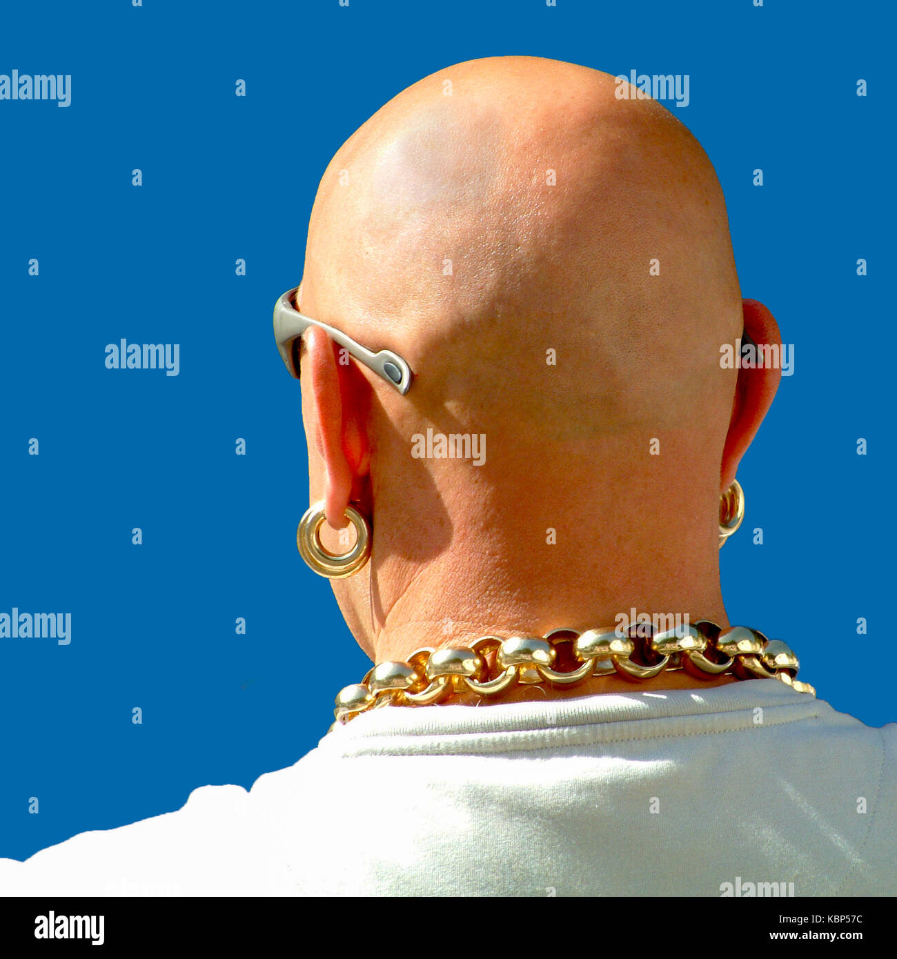 Head shot of shaven-headed man from behind wearing sunglasses , heavy gold earrings and a very heavy gold chain against a clear blue sky. Stock Photo