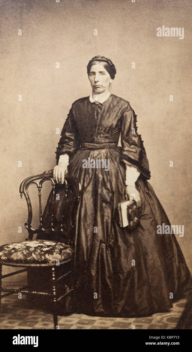 American archive monochrome studio portrait photograph of older woman with stern expression wearing a dark dress standing next to upholstered dining room chair, named as Matilda Gage, taken in late 19th century by Webster and Battey Photographers, Binghamton, NY, USA Stock Photo
