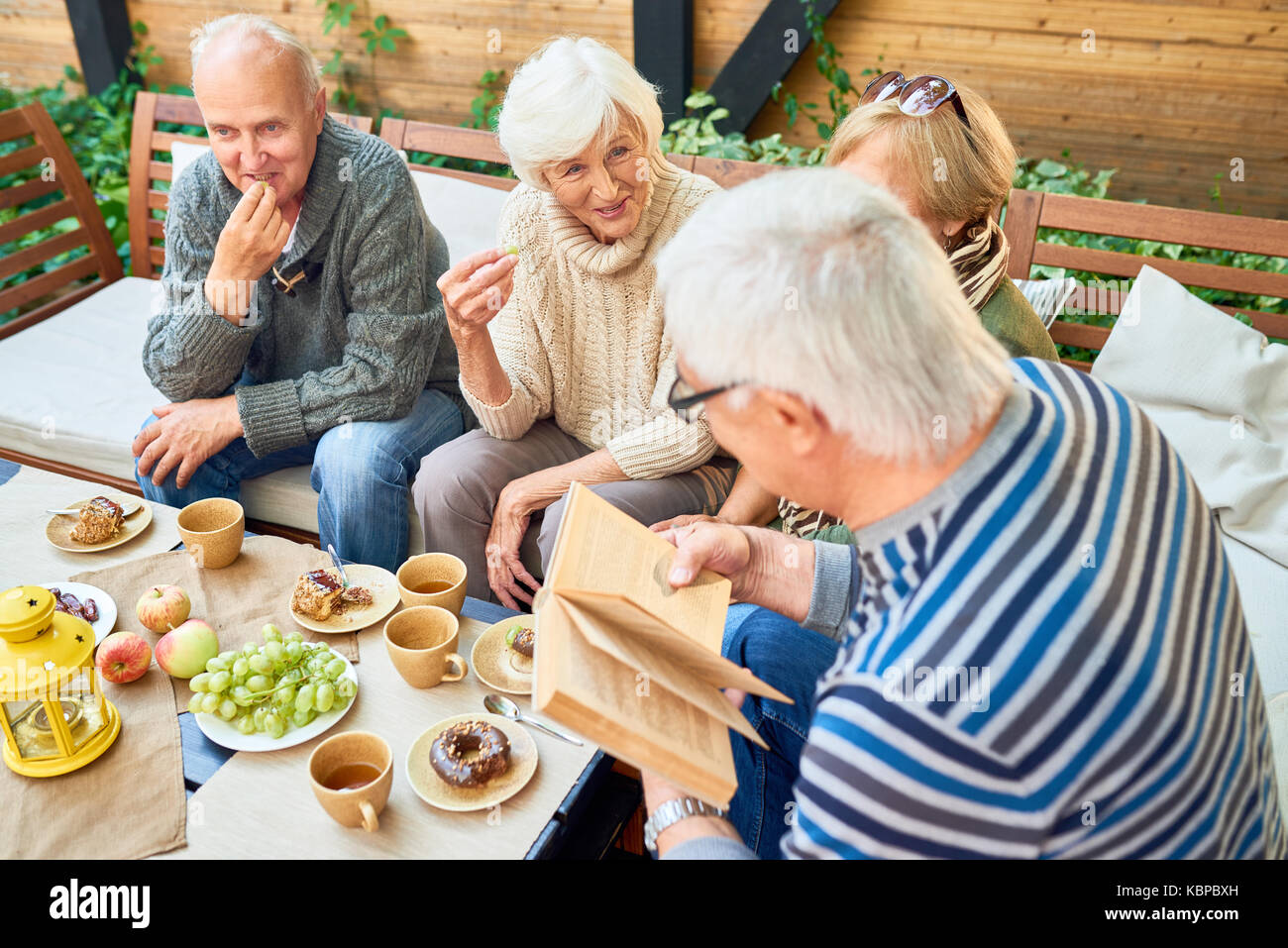 Group of joyful senior friends having fun outdoors: they enjoying delicious pastry and fragrant coffee while elderly man in striped sweater reading th Stock Photo