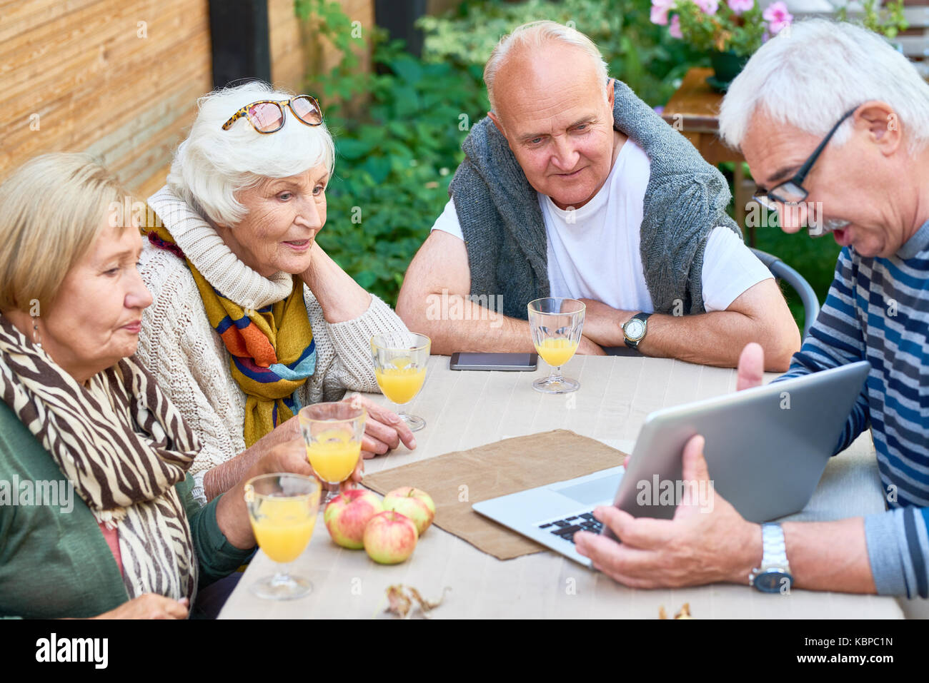 Two senior couples spending weekend together at backyard: they looking through photos on laptop and enjoying freshly squeezed juice Stock Photo