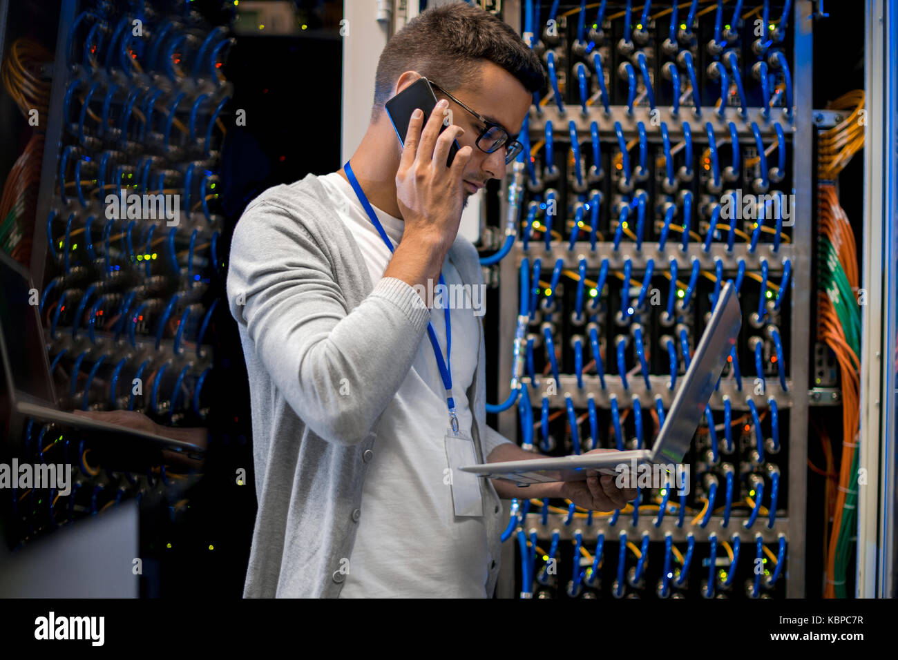 Side view  portrait of young man speaking by phone and using laptop against background of blade servers in supercomputer Stock Photo