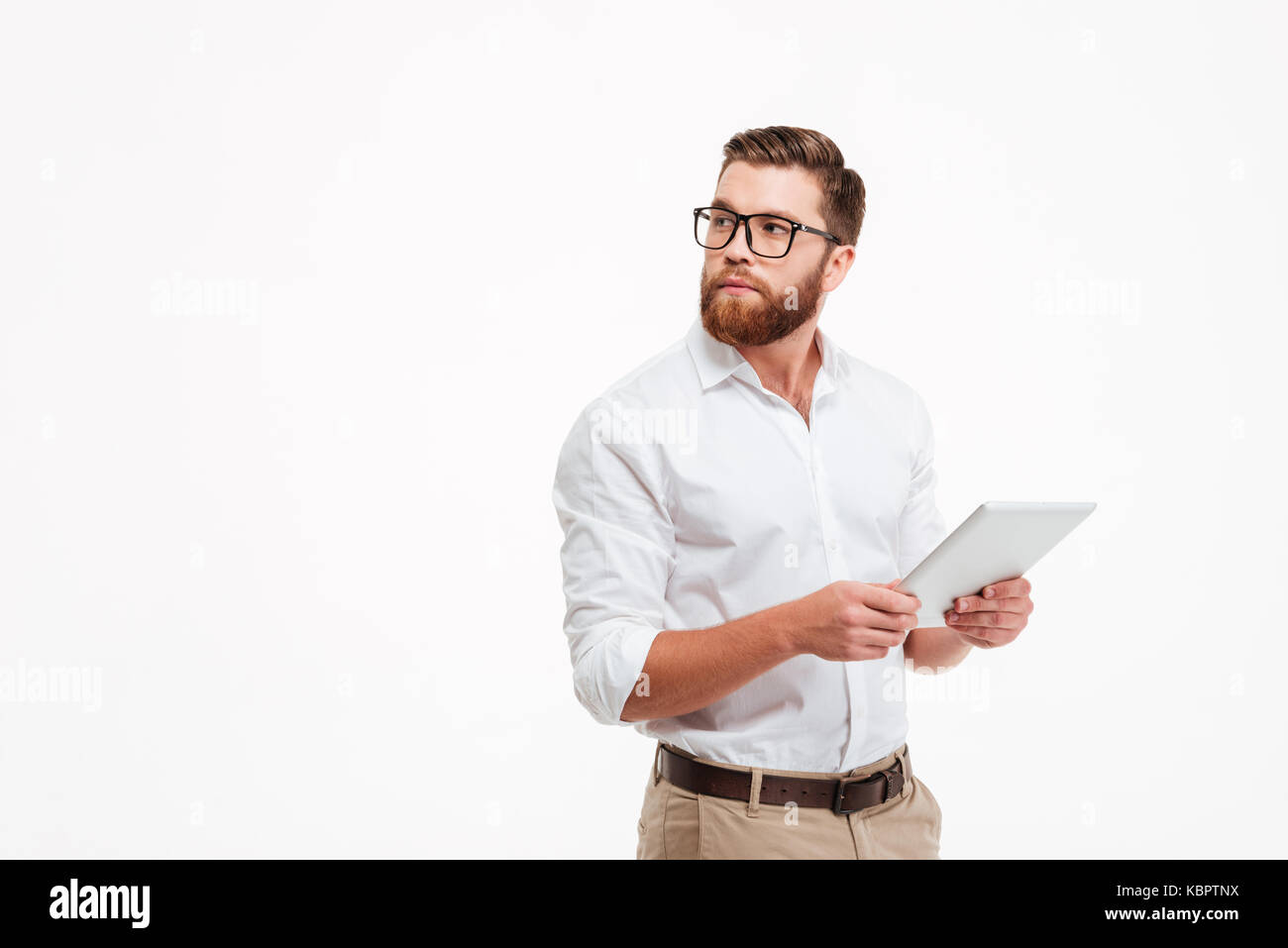 Picture of serious young bearded man standing near copyspace over white wall background isolated. Looking aside using tablet computer. Stock Photo