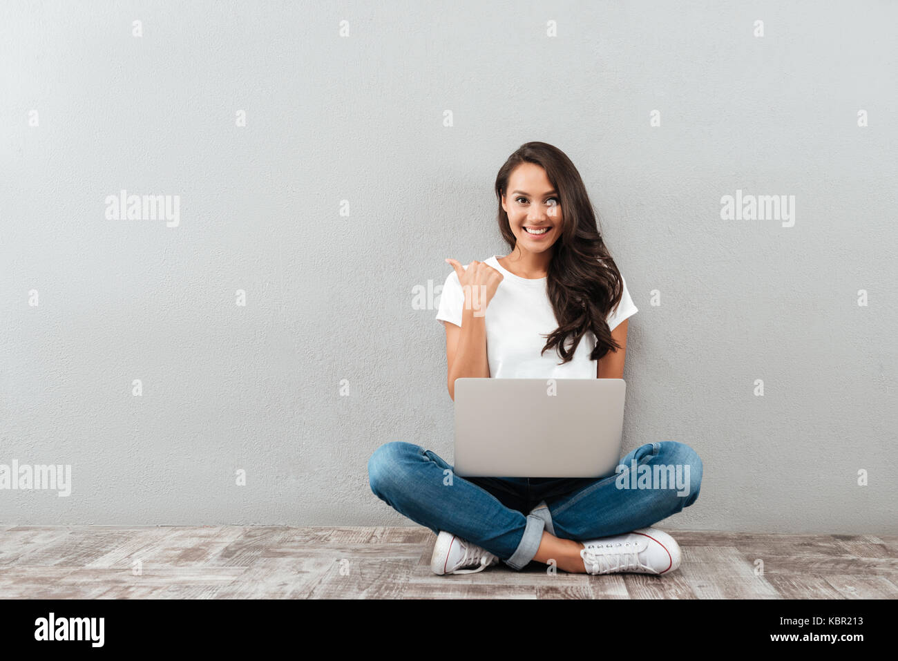 Happy smiling asian woman working on laptop computer while sitting on the floor with legs crossed and pointing finger away isolated over gray backgrou Stock Photo