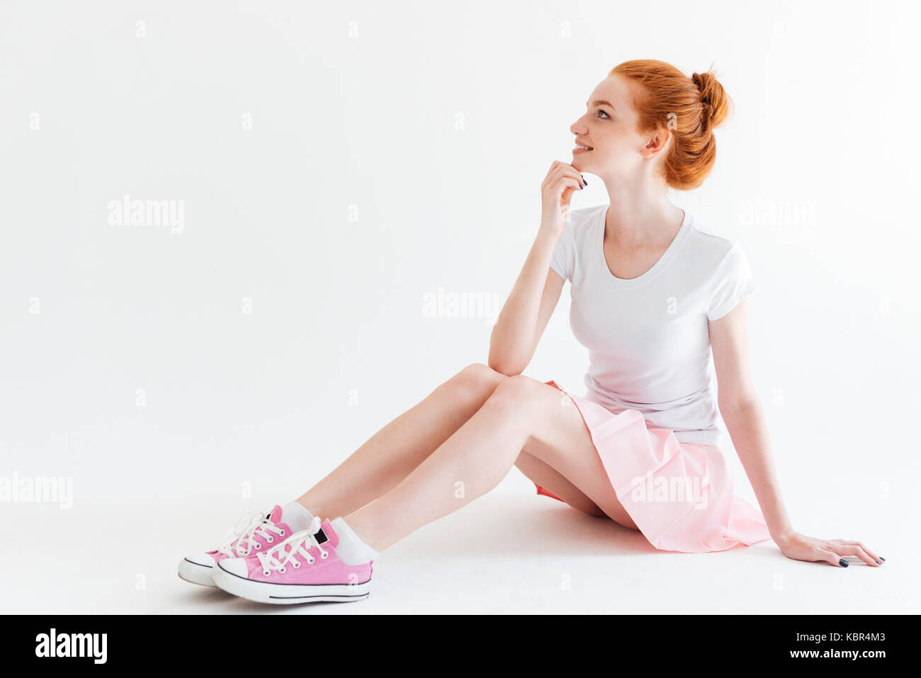 Smiling ginger girl sitting on the floor and looking away over white ...