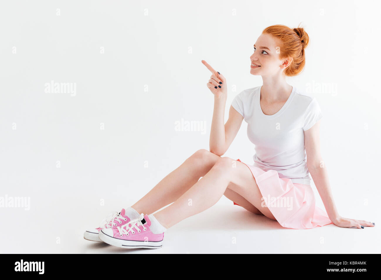 Smiling ginger girl sitting on the floor and pointing away over white ...