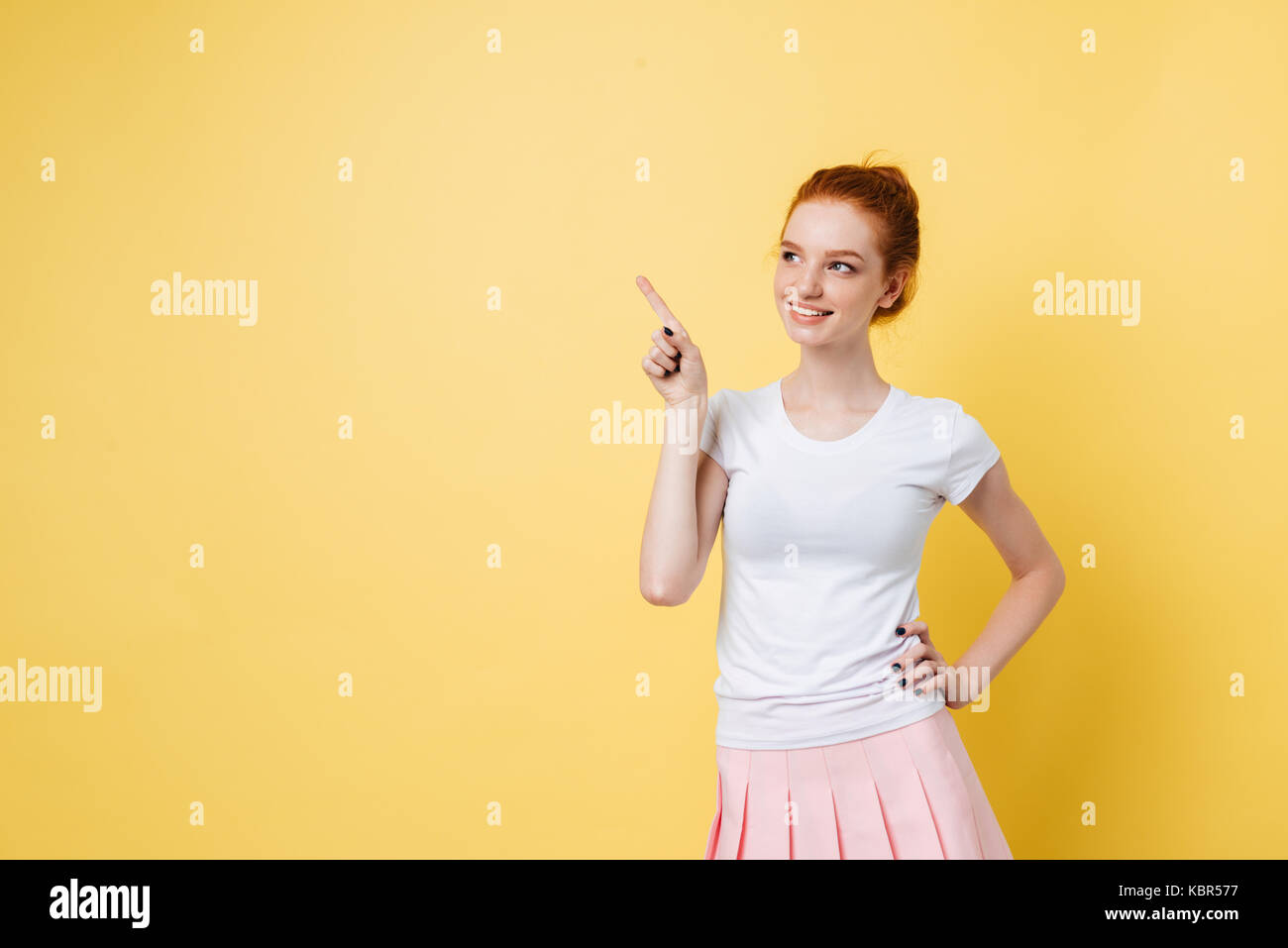 Carefree ginger girl pointing away and holding arm on hip over yellow ...