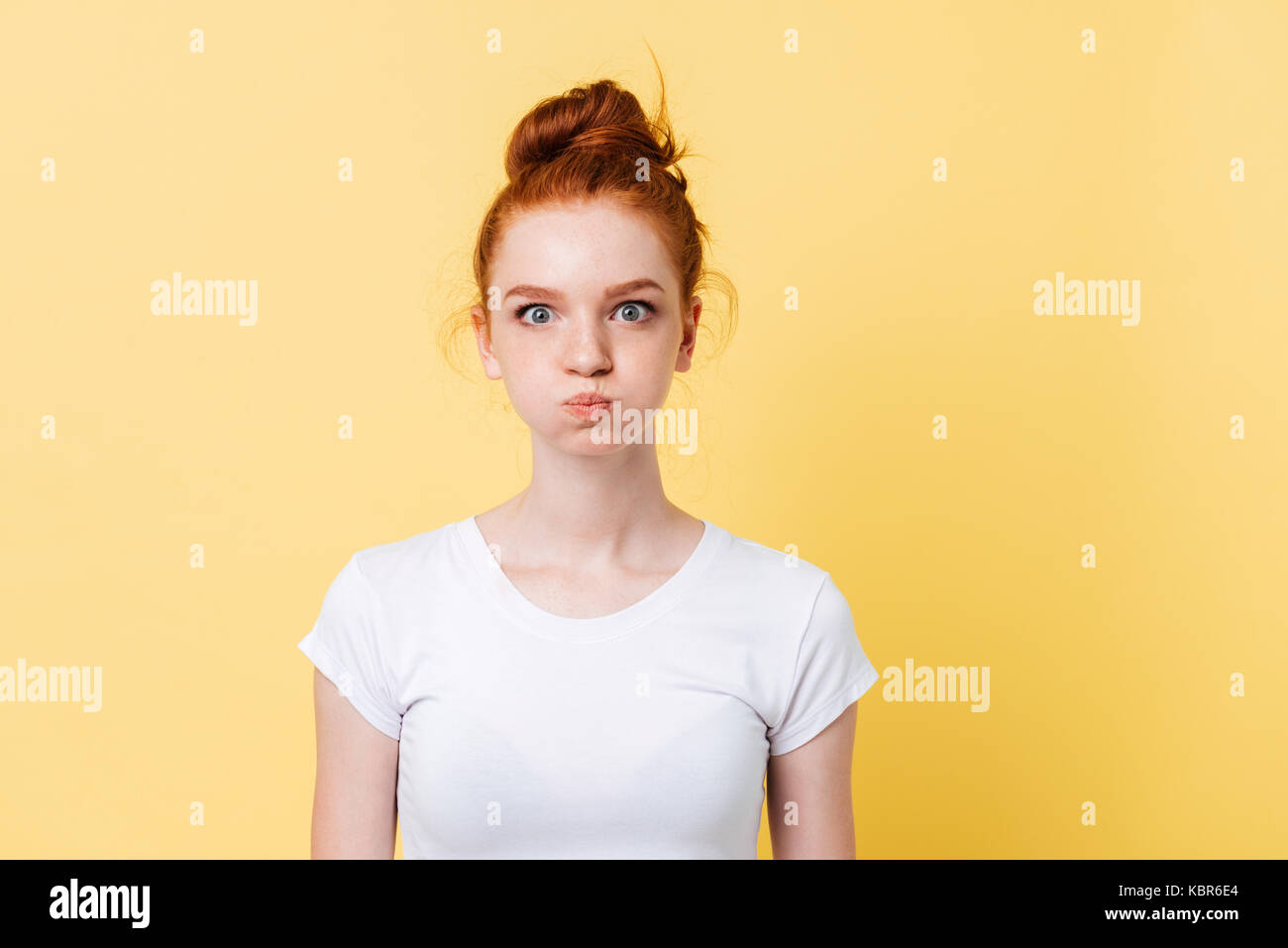 Pretty ginger woman in t-shirt with puffed cheeks looking at the camera over yellow background Stock Photo