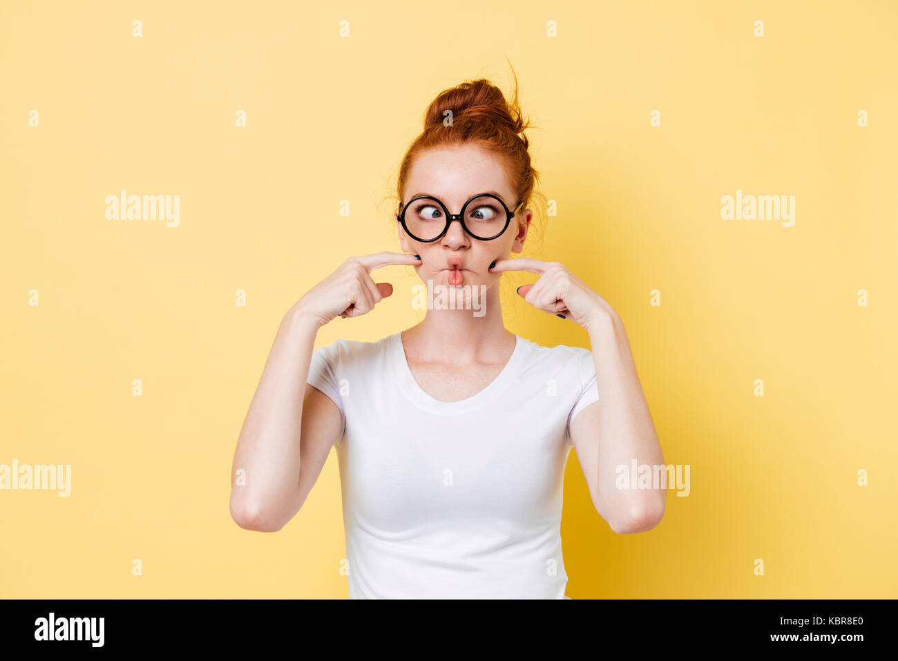 Funny ginger woman in eyeglasses puffed-off cheeks over yellow background Stock Photo