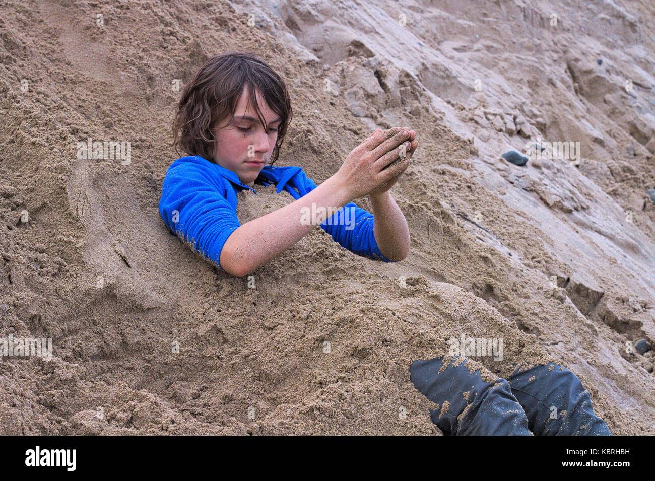 Young teenage boy covering himself in sand while fully dressed, winter at Marengo beach, Vic, Australia. Stock Photo
