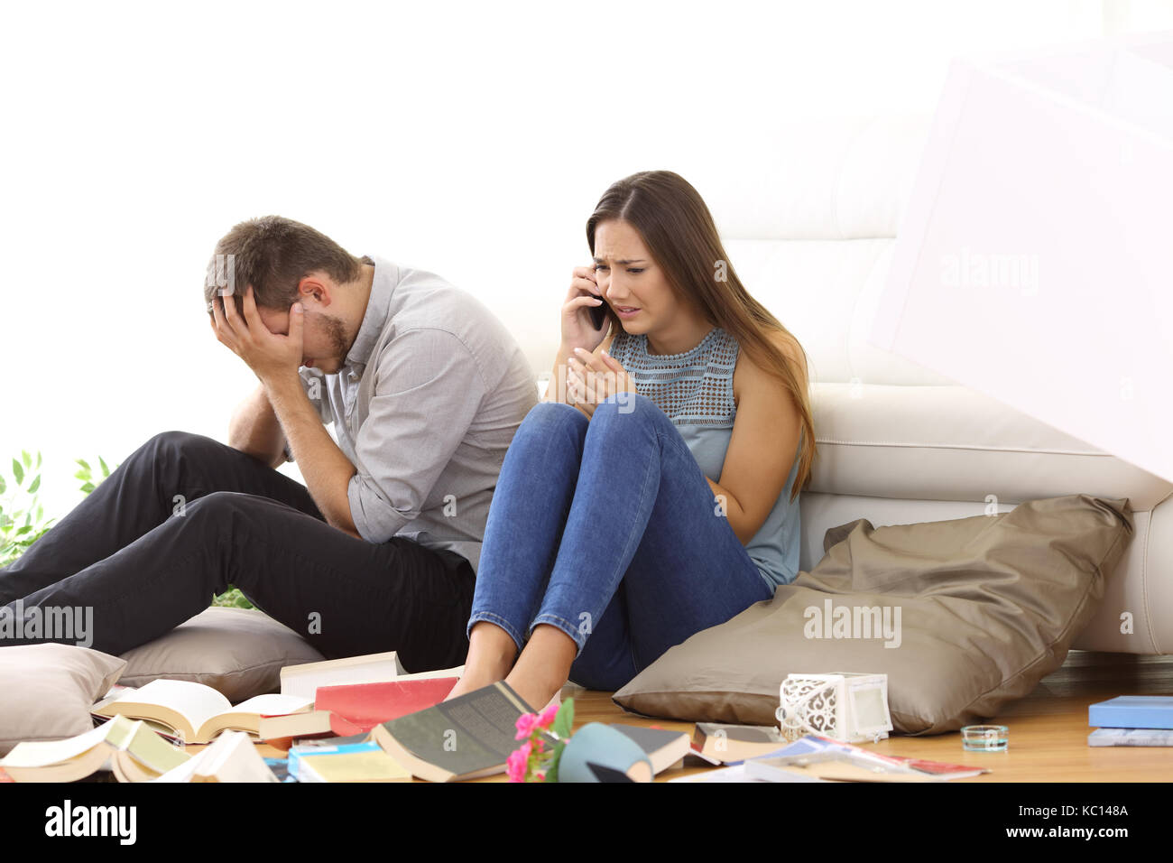 Desperate couple calling police after a robbery sitting on the floor of a messy living room at home Stock Photo