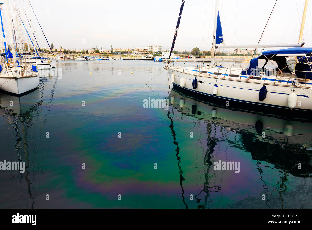 Yachts moored in Zygi harbour with fuel pollution casting a rainbow sheen on the water surface. Stock Photo