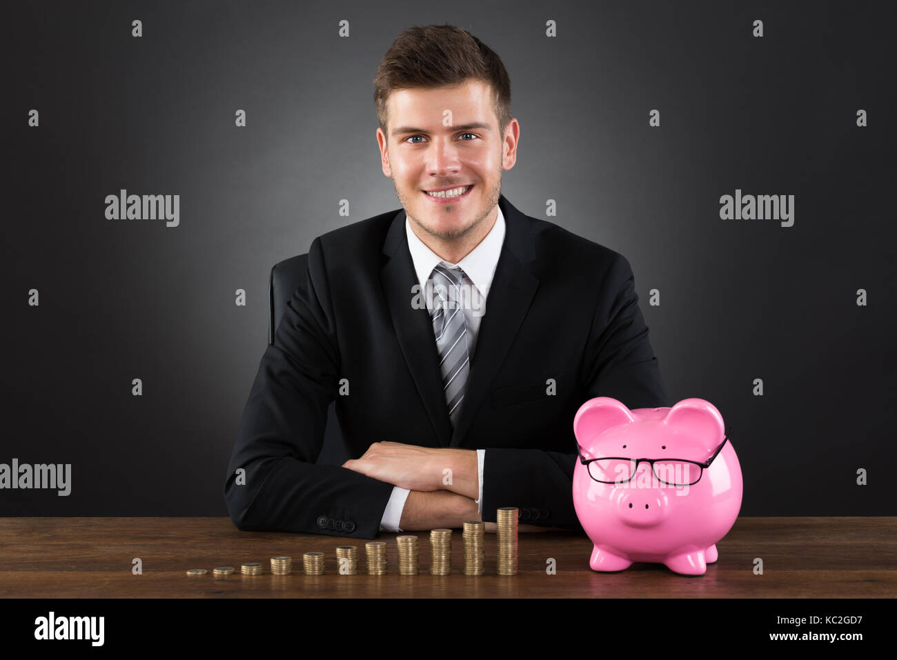 Young Businessman With Stack Of Coins And Piggybank At Desk Stock Photo