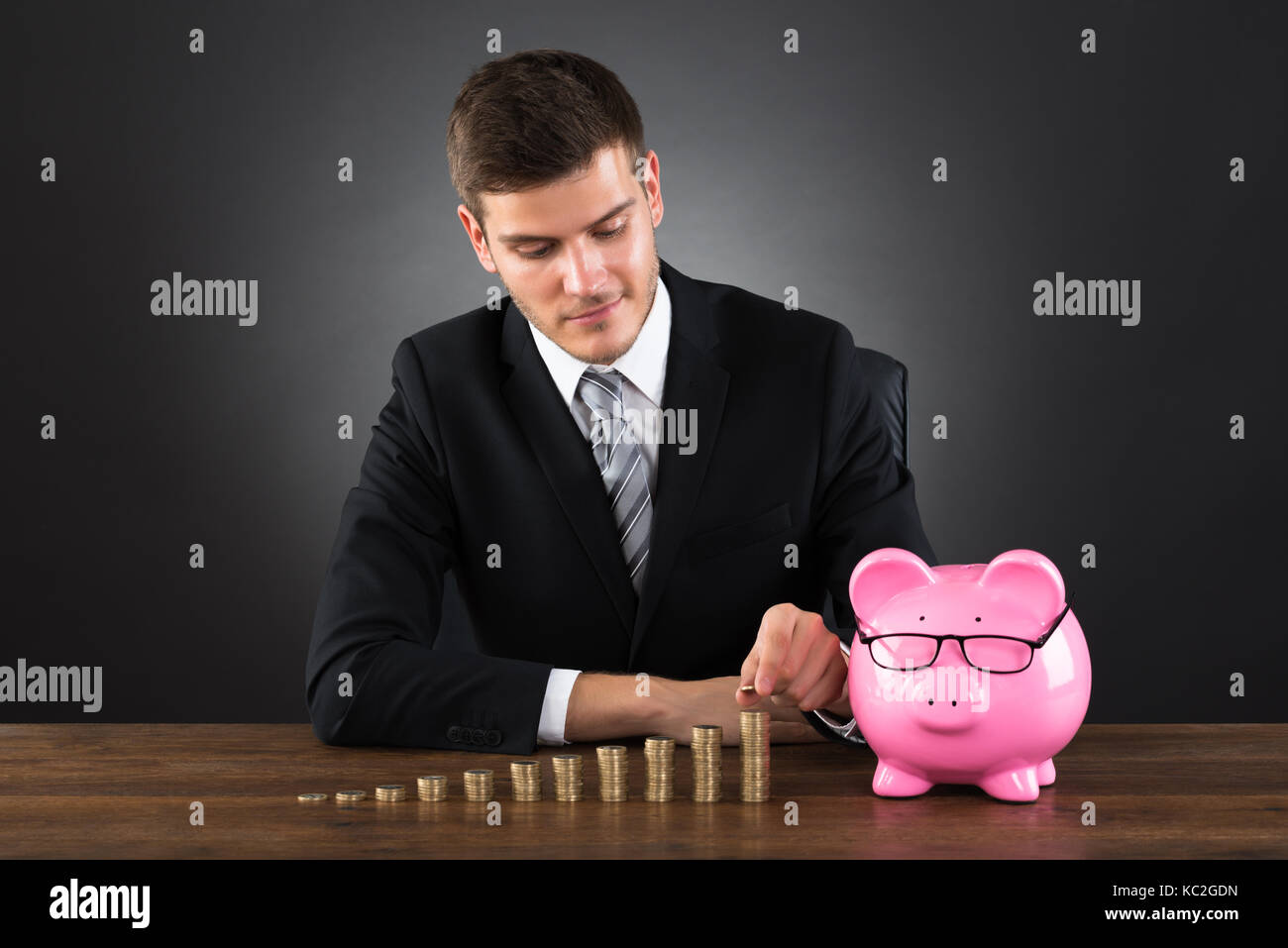 Portrait Of Young Businessman With Piggybank Stacking Coins On Desk Stock Photo