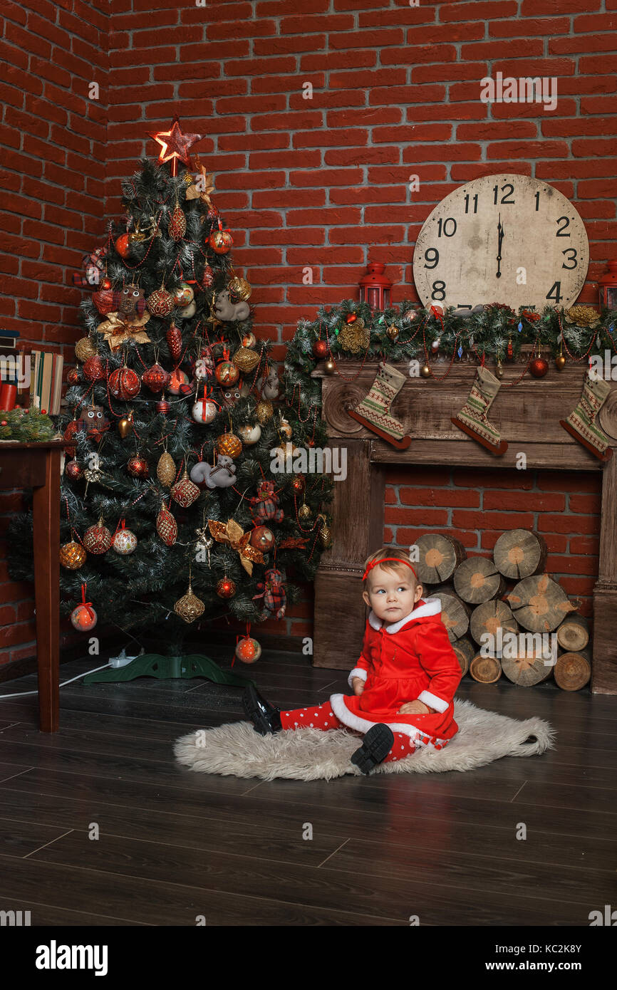 Baby girl dressed as Santa Claus sits near a Christmas tree Stock Photo