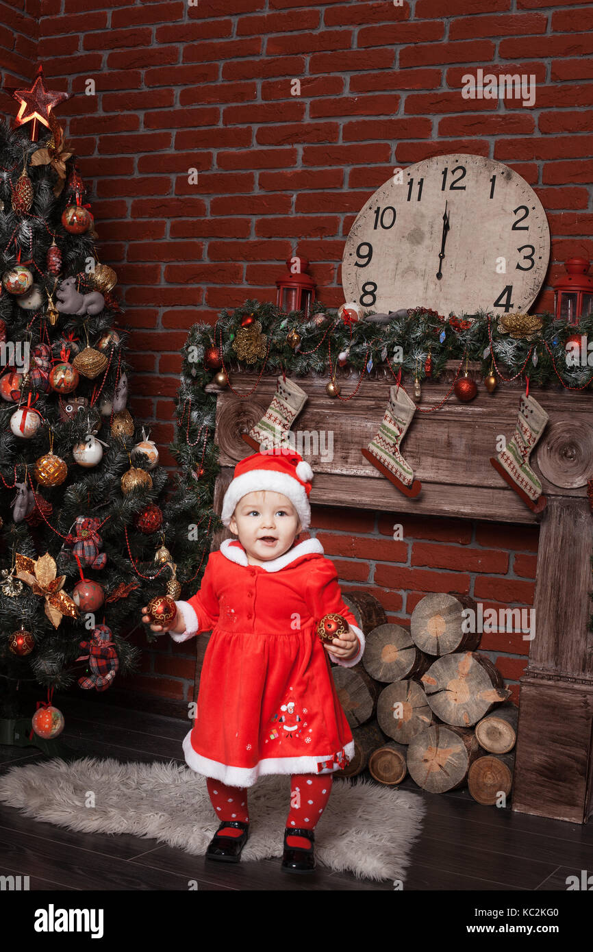 Baby girl dressed as Santa Claus stands near a Christmas tree and holding Christmas balls Stock Photo