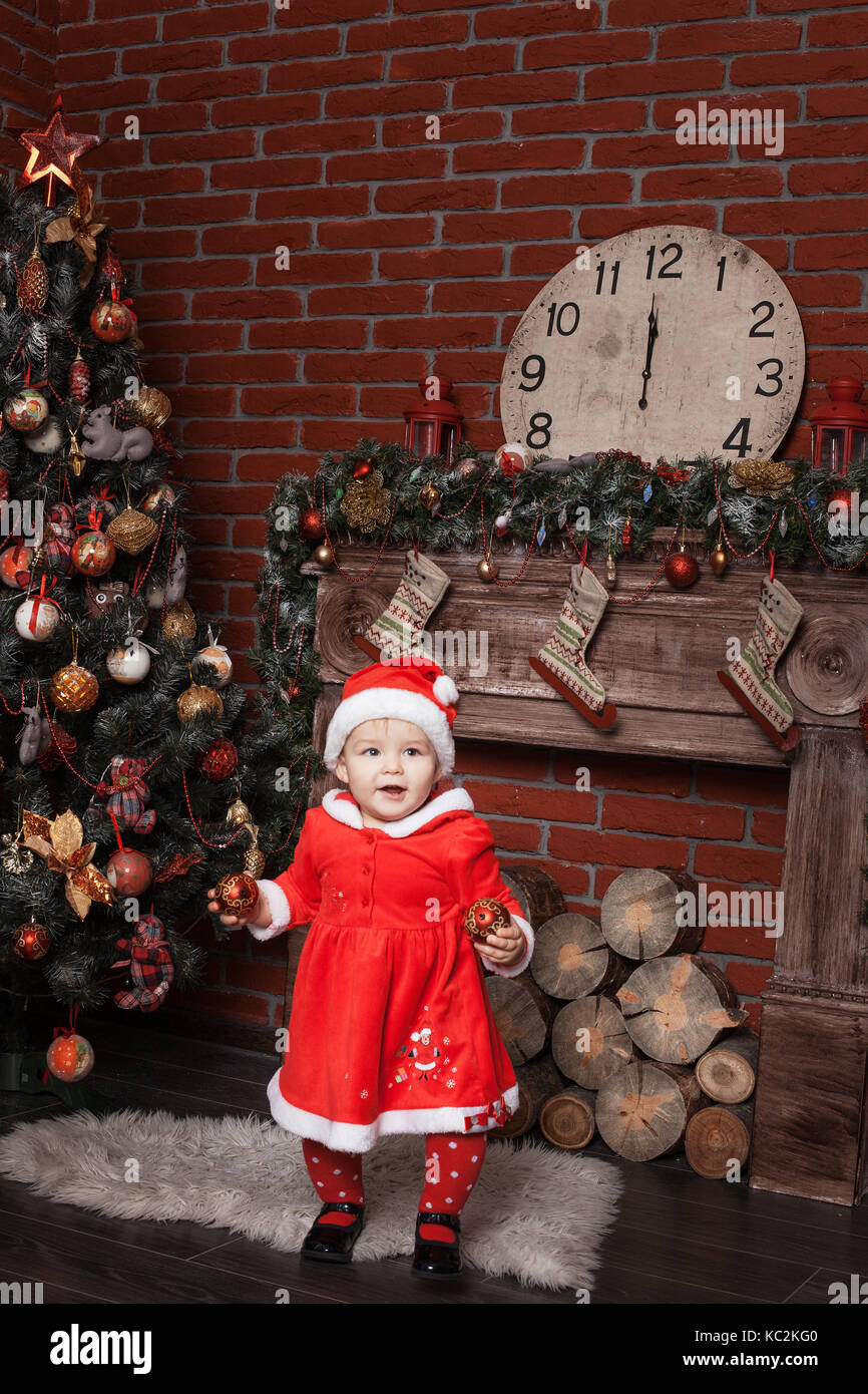 Baby girl dressed as Santa Claus stands near a Christmas tree and