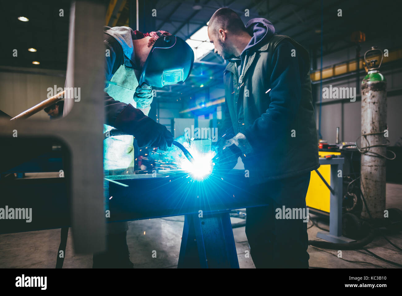 Industrial Welder With Torch and Protective Helmet in big hall welding metal profiles Stock Photo
