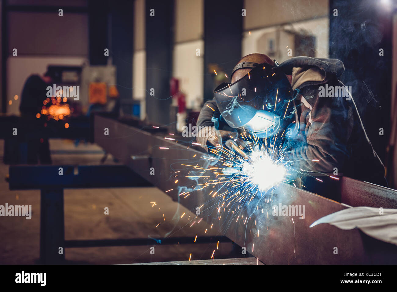 Industrial Welder With Torch and Protective Helmet in big hall welding metal profiles Stock Photo