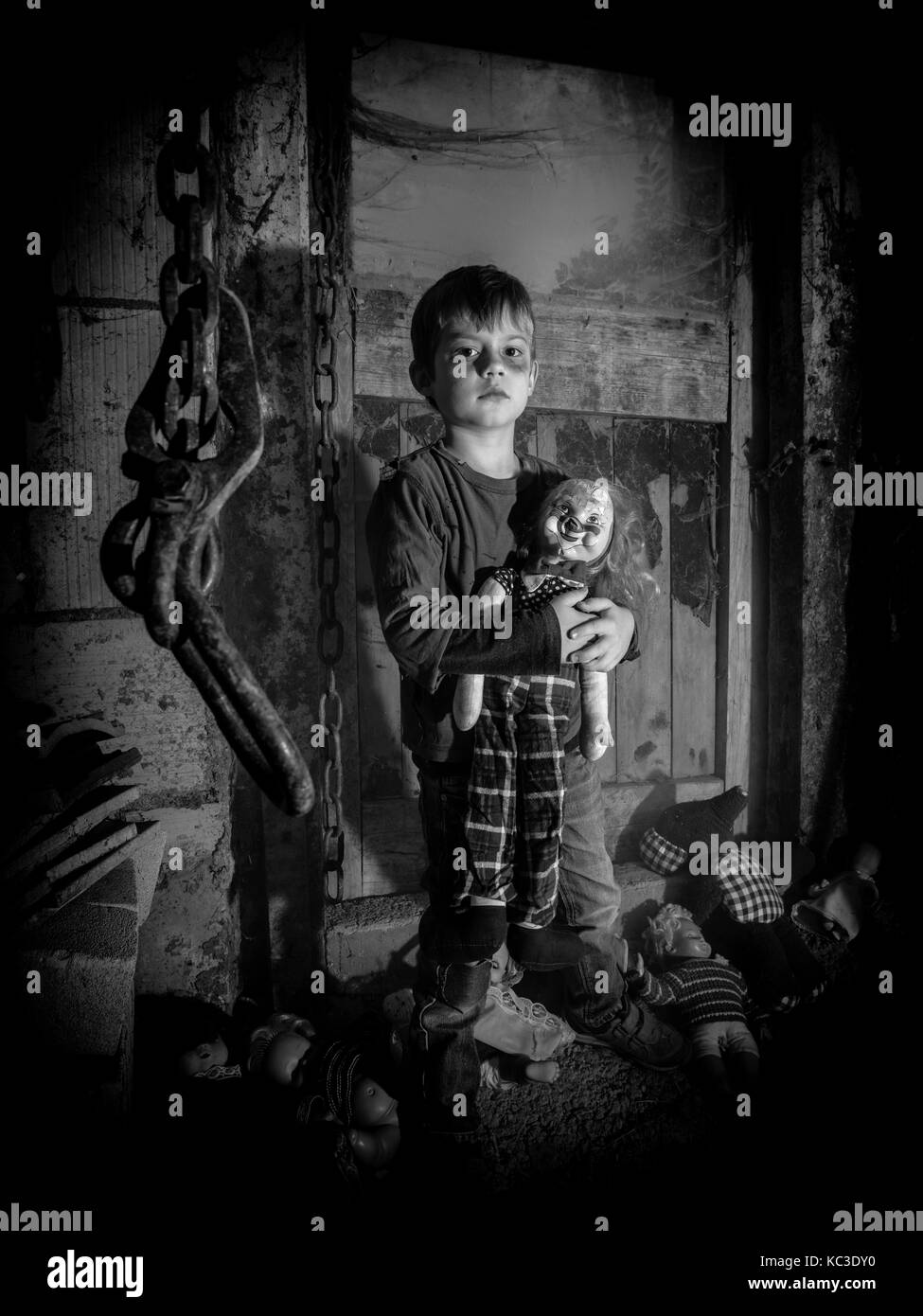 Photo of a creepy young boy holding an old clown doll in an old barn covered in spiderwebs and dust. Stock Photo